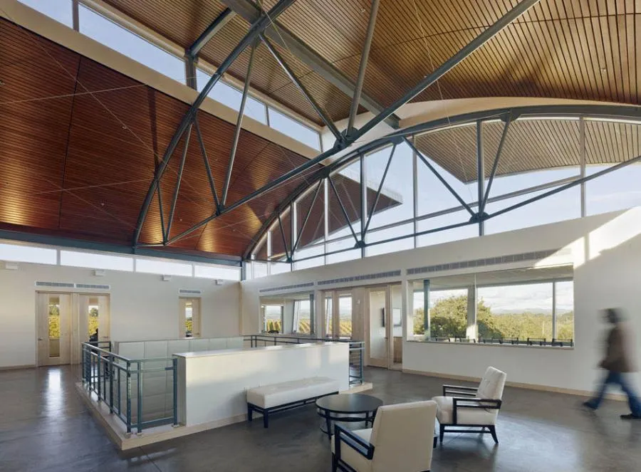  Spacious office interior with high, curved wood-paneled ceilings, large windows, and minimalistic furniture, including chairs and a table. A blurred figure walks across the room.