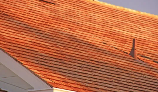  Close-up of a rooftop with neatly arranged reddish-brown shingles in warm lighting, including a vent pipe, illustrating roofing craftsmanship.