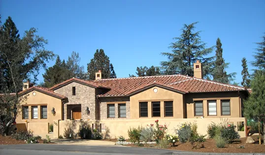  A stucco house with a tile roof surrounded by trees and shrubs, set under a clear blue sky.