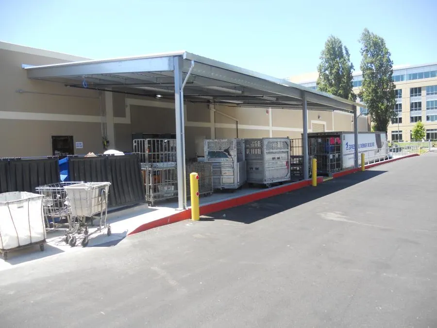Outdoor loading area with metal carts and covered storage, next to a building with trees in the background.