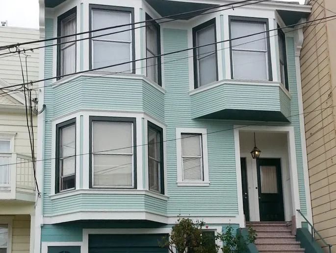 A two-story house with a fresh light blue paint job, featuring bay windows and a small front staircase leading to a black door.