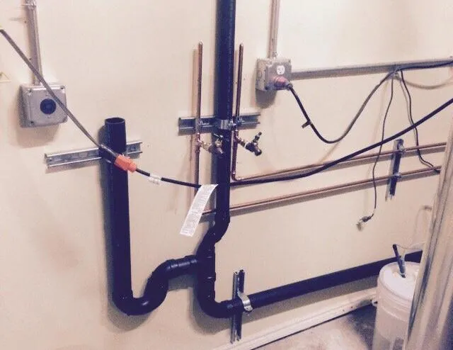  Plumbing installation on a wall featuring various black pipes, copper lines, and electrical connections, with a bucket nearby.