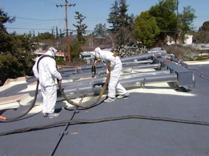 Two workers in protective suits applying material on a flat roof with hoses, surrounded by trees and utility poles.