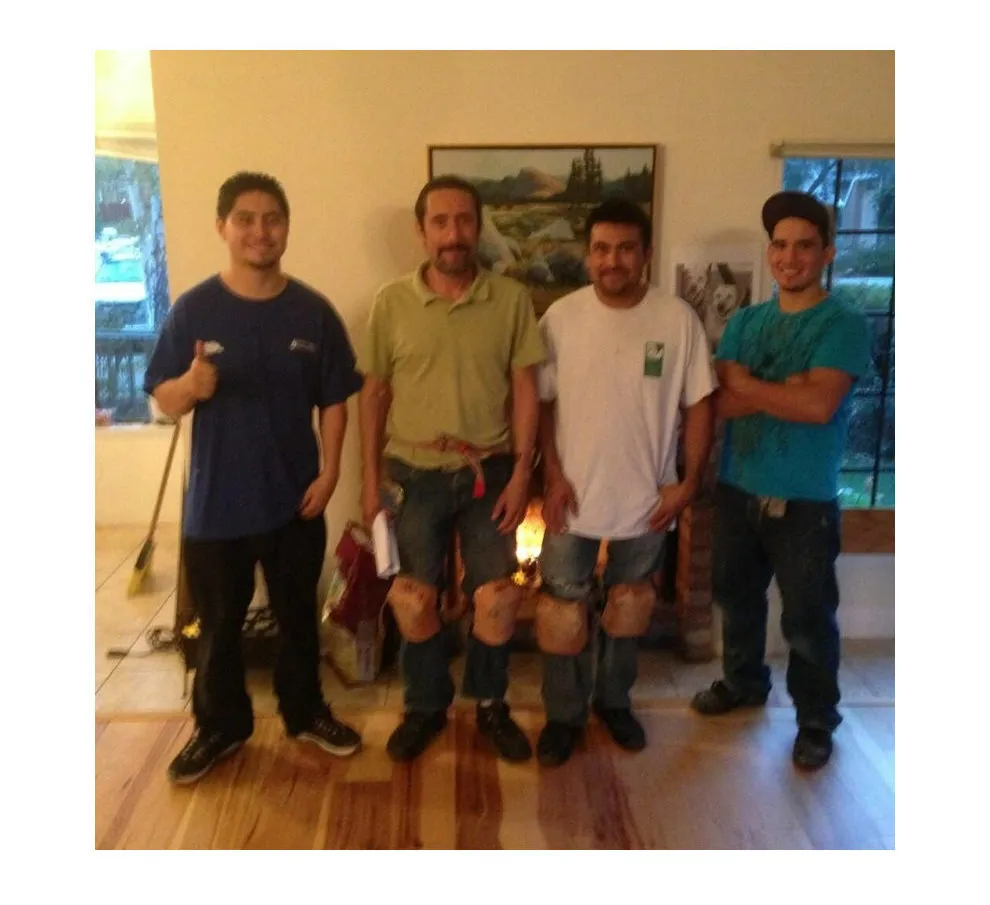  Four smiling workers standing on a newly installed hardwood floor, with tools and a broom visible in the background. A painting and windows are on the walls.