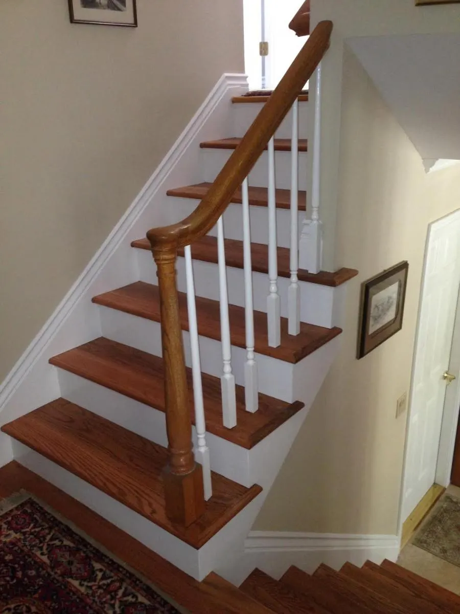 Wooden staircase with a polished finish, featuring white spindles and a wooden handrail, in a carpeted area with framed artwork on the walls.