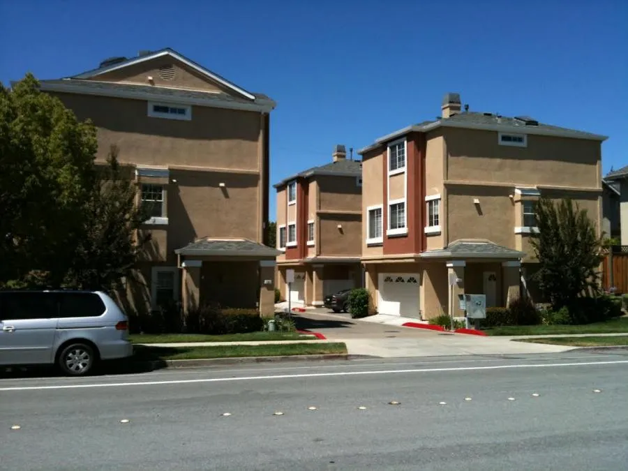 View of two beige and brown townhomes with garages, surrounded by trees and a parked vehicle on a sunny day.