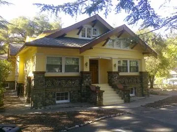 Yellow house with stone accents, front steps, surrounded by trees, under clear sky.