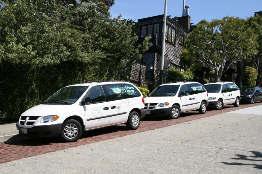  Three white SonoMarin Cleaning Services vans parked in a row on a residential street beside trees and houses.