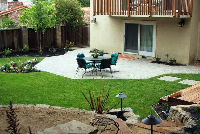 A landscaped backyard with a patio, outdoor table and chairs, green lawn, flower beds, and a sliding glass door.