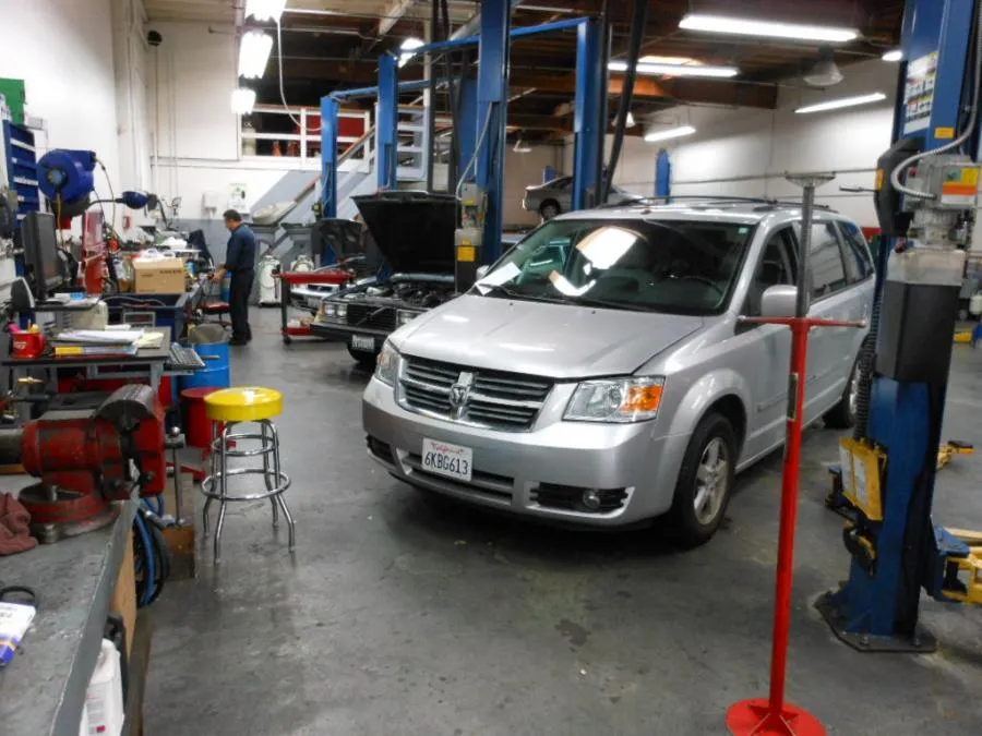  A silver van is parked in a busy auto repair shop with various tools and equipment. A mechanic is seen working in the background near a vehicle with an open hood.
