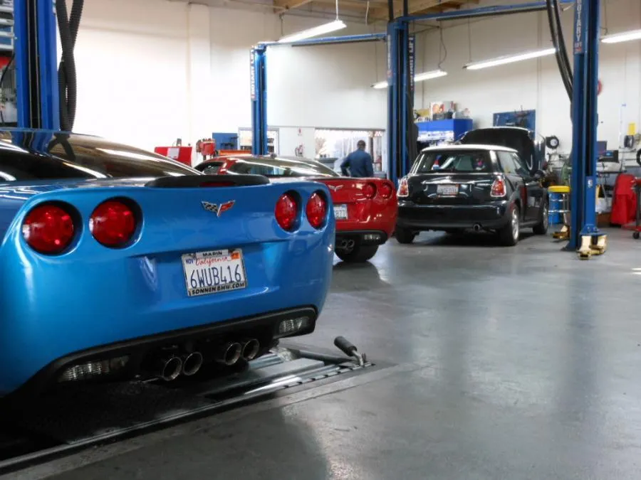 Automotive workshop with cars, including a blue Corvette, under repair. Technician visible in the background.
