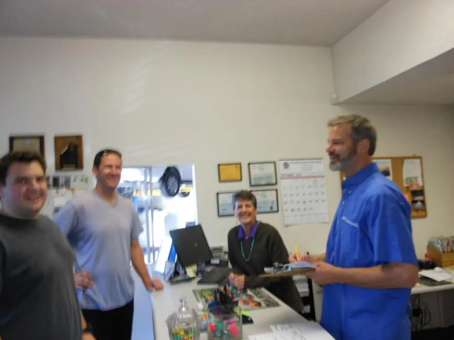 Four people in an office, standing near a desk with computers and office supplies. Walls have certificates and a calendar.