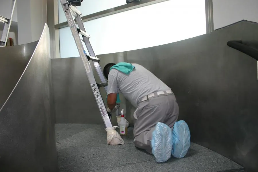  Person kneeling and cleaning a floor near a ladder, with cleaning supplies beside them and wearing shoe covers.