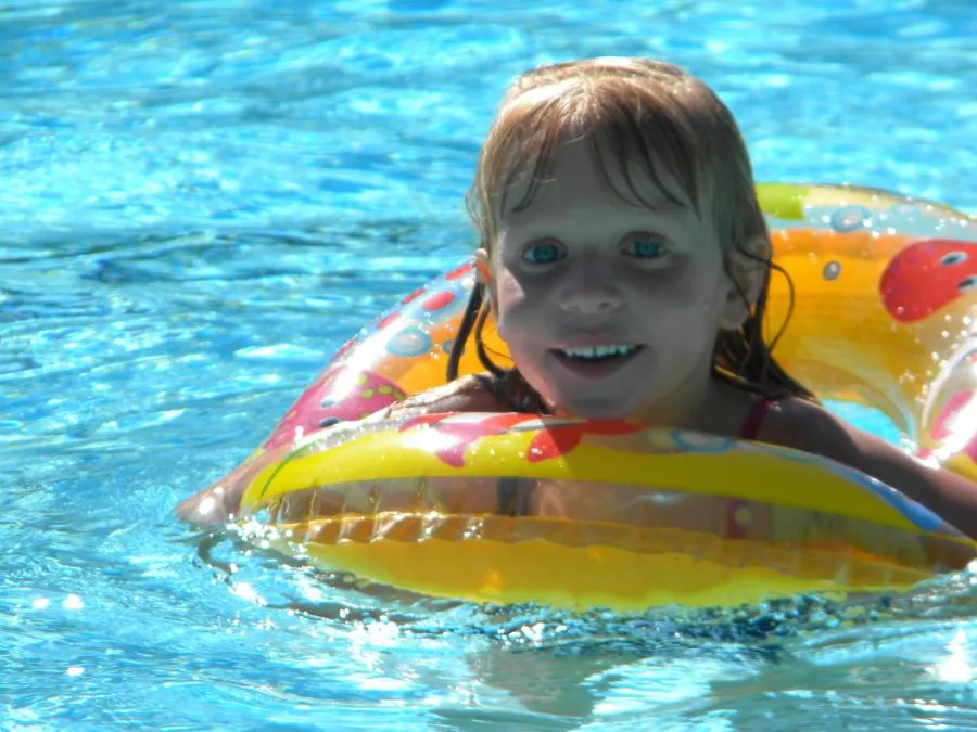  A child in a pool smiles while using a colorful inflatable ring. The water is clear and sunlit.