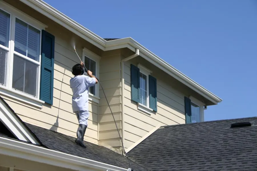  Person on a roof power washing the exterior of a beige house with blue shutters under a clear blue sky.