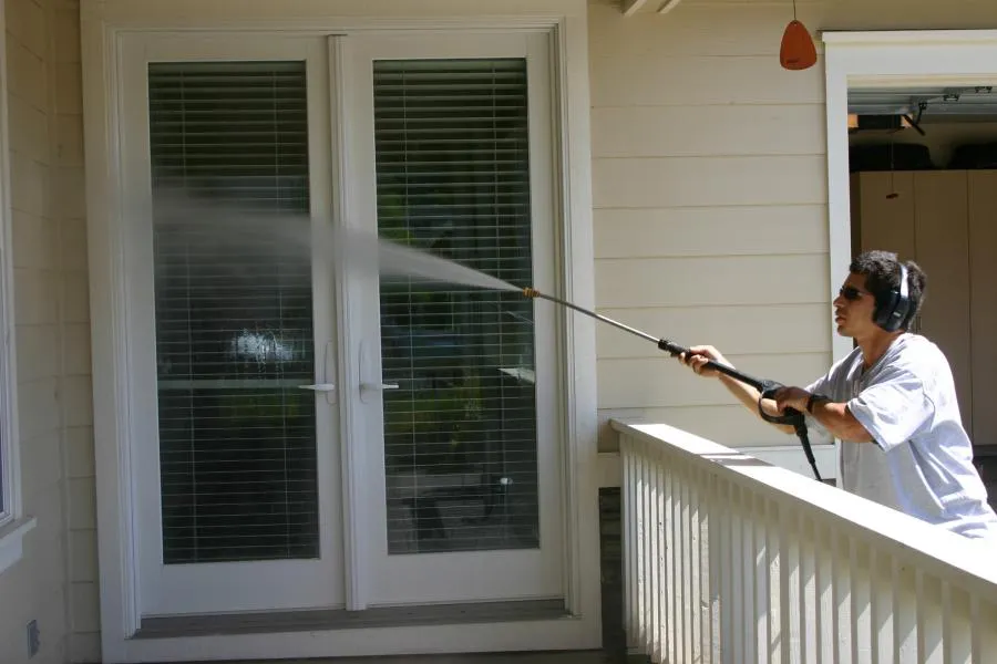  A person using a pressure washer to clean glass doors, wearing headphones for protection, on a porch with a railing.