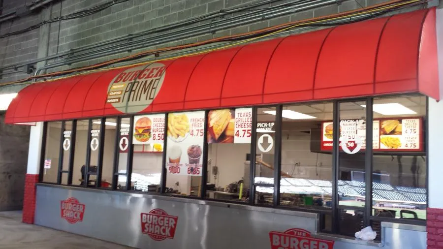 Fast-food counter with a red awning, signs for burgers and fries, and a pickup window in an industrial setting.