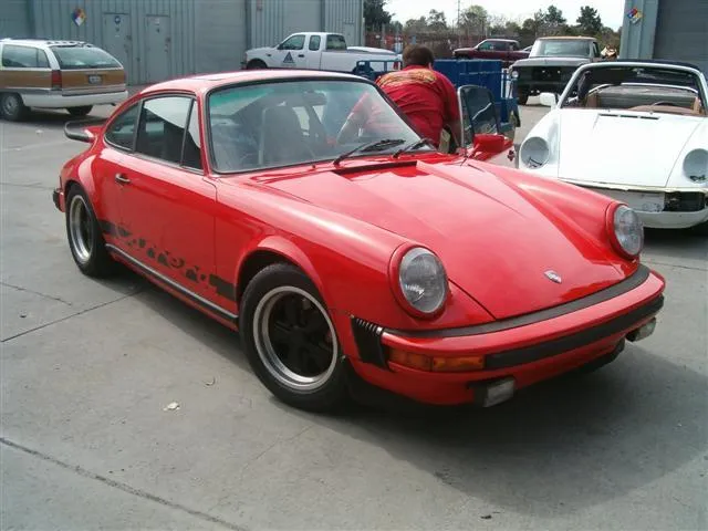 A red classic car is parked outdoors near a person working on another vehicle, with more cars and a building in the background.