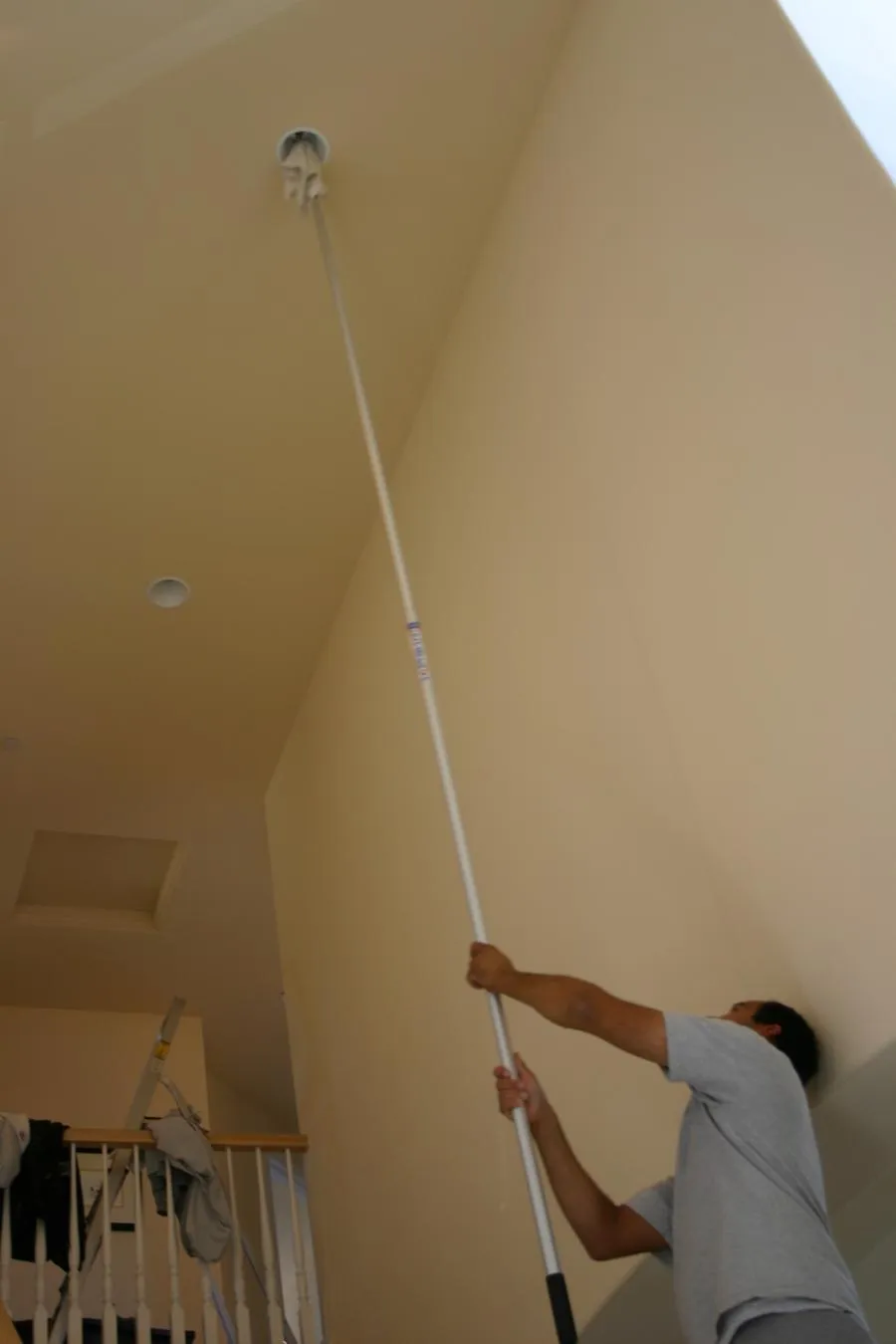  A person uses an extendable pole to clean or change a ceiling light in a residential interior with high ceilings. A ladder and some cloths are visible in the background.
