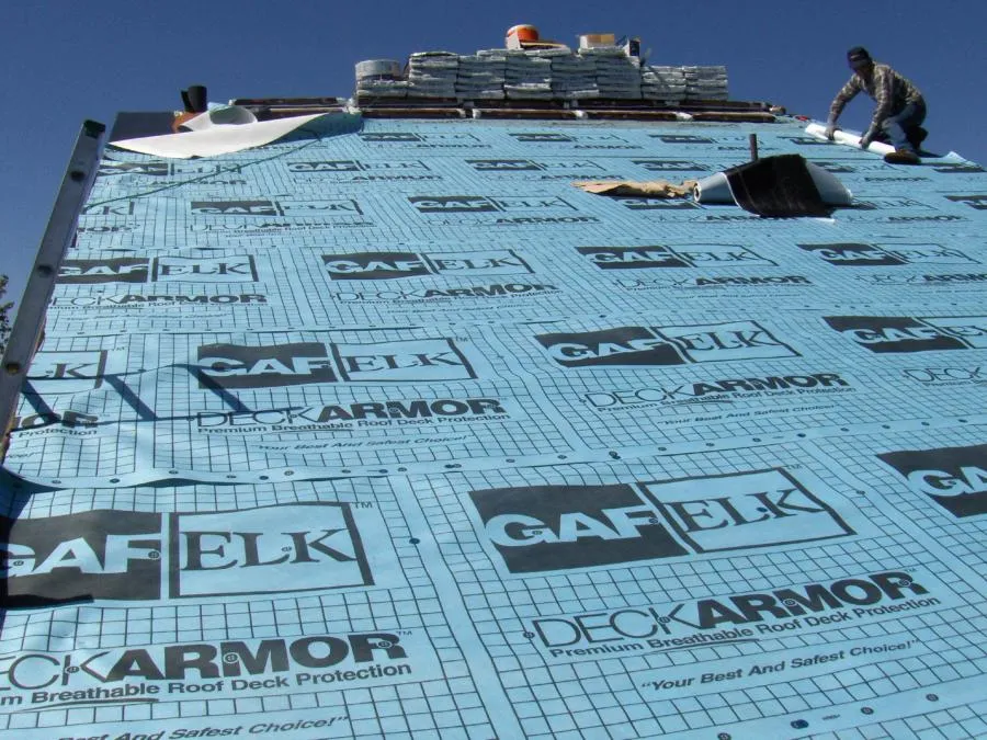  Roofer working on a roof covered with blue GAF Deck Armor underlayment, with stacks of roofing material at the top.