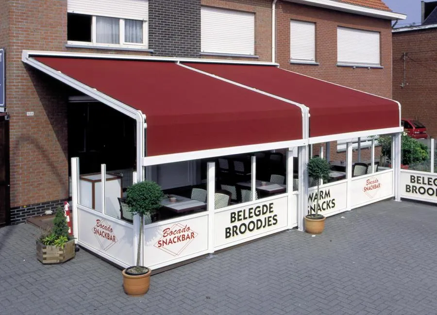  Outdoor seating area with red awnings, white barriers reading "Bocado Snackbar" and "Belegde Broodjes". Potted plants and tables with chairs are visible.