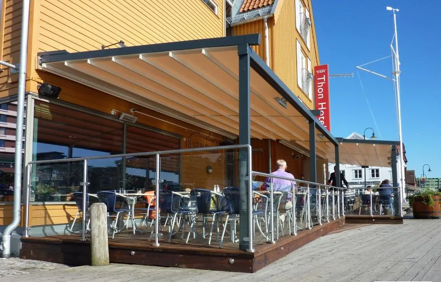 Outdoor café terrace with metal and glass railing, featuring a retractable awning attached to a yellow building. Tables and chairs are arranged on the wooden deck under a clear blue sky.