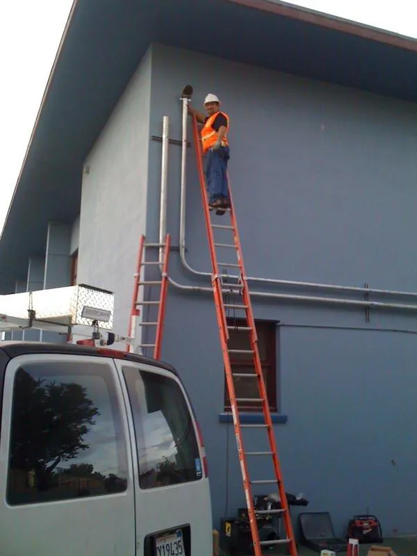  A technician in a safety vest and hard hat works on an electrical fixture atop a ladder against a building's wall, with a van and equipment nearby.