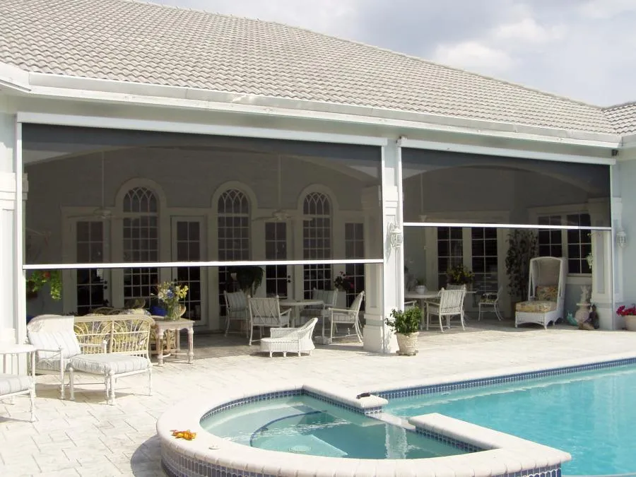  Outdoor patio with retractable sunshades, wicker furniture, and a pool in the foreground. White arches and glass doors in the background.