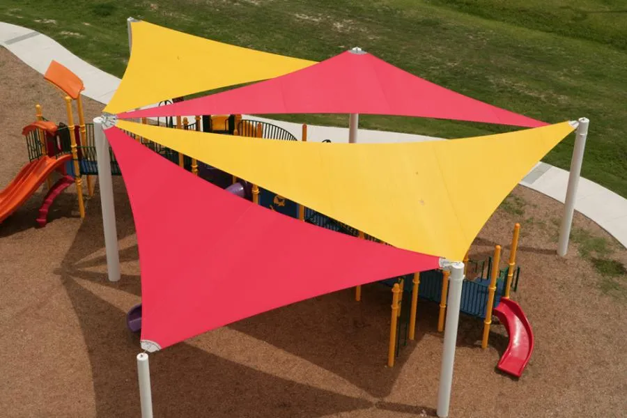  Aerial view of a playground with three large triangular sunshades in red and yellow, supported by white poles.