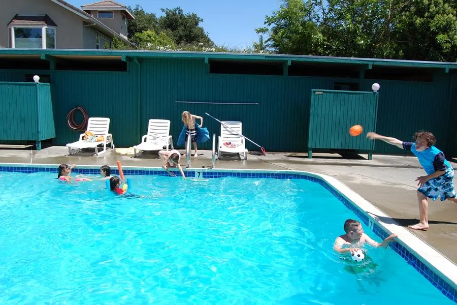  Kids playing in a swimming pool, with others on the deck. A child throws an orange ball, while another swims. Lounge chairs and a fence are in the background.