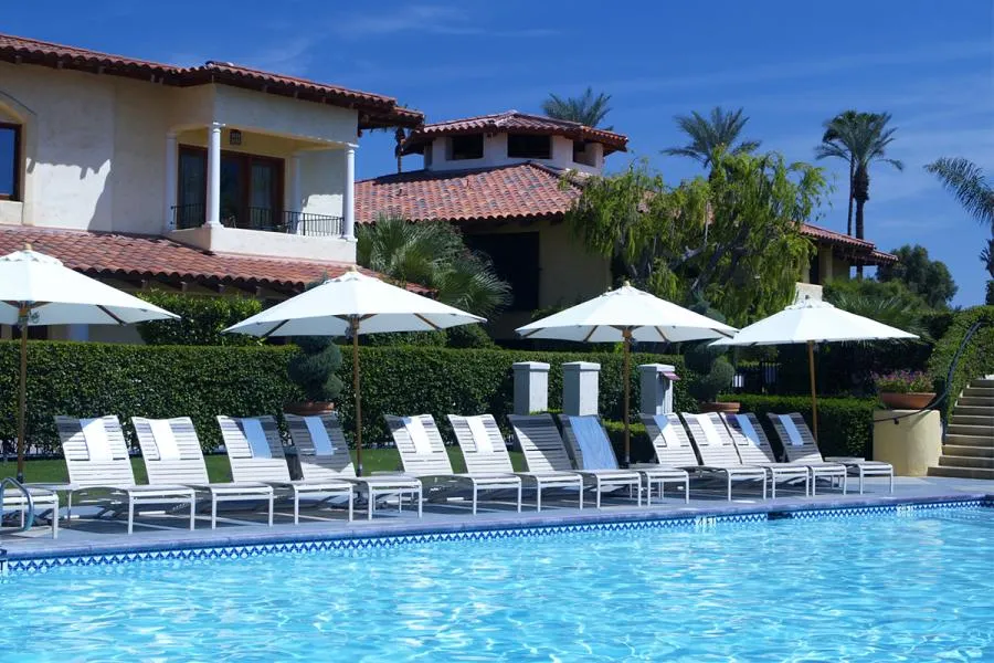 Outdoor pool area with lounge chairs and umbrellas, surrounded by a lush garden and a building with terracotta roofs.