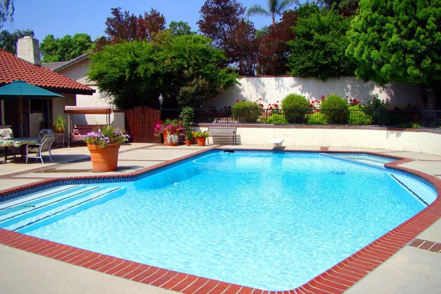 A clear blue swimming pool with brick edging, surrounded by patio furniture, potted plants, and a lush garden backdrop.