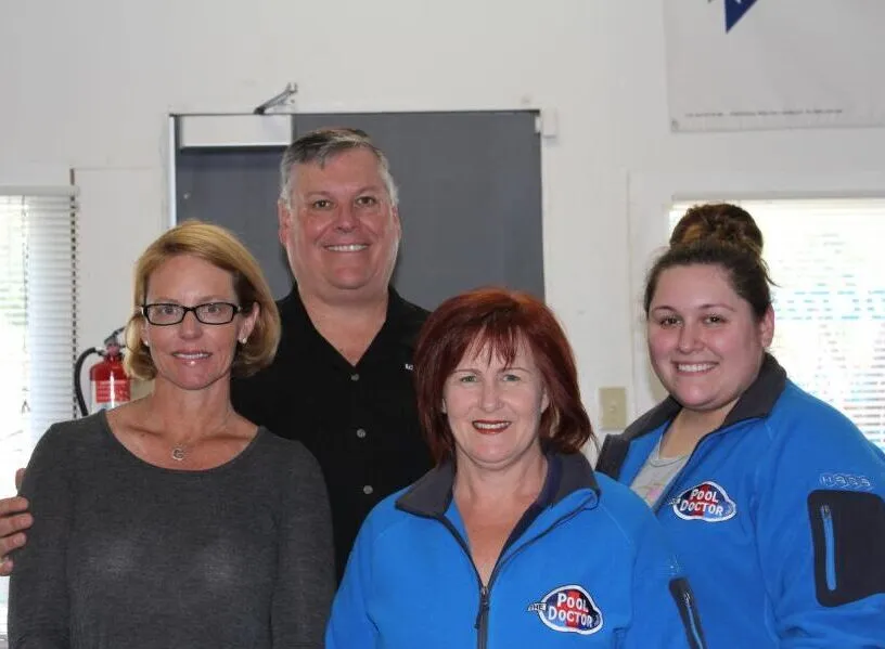  Four people posing together indoors, with two wearing The Pool Doctor jackets and a sign visible in the background.