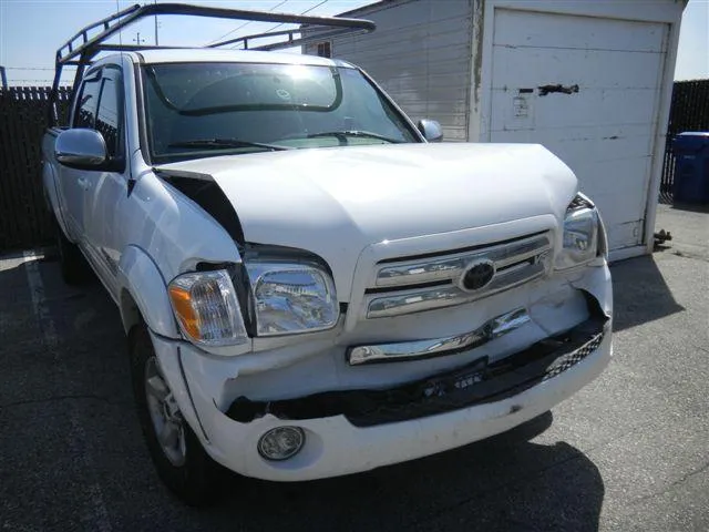  Damaged white truck with crumpled front bumper and hood, parked outside near a garage door in a sunlit area.