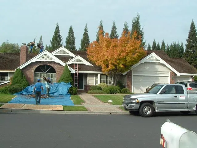  Workers on a roof, ladder propped against a house, tarps covering the ground, and a company truck parked in the driveway.