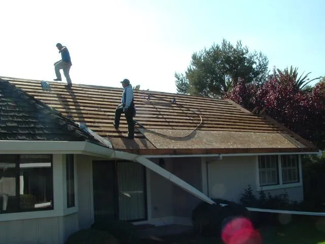  Two workers standing on a residential roof under renovation, with tools and equipment visible against a backdrop of trees.