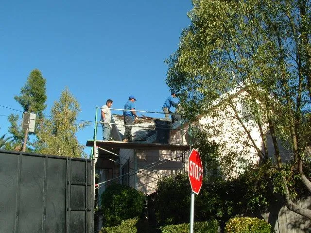  Workers on scaffolding replace roofing shingles on a residential home, surrounded by trees and clear skies.