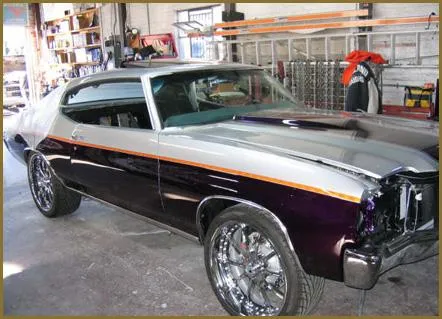  A classic car with shiny custom paint and large chrome wheels in an auto shop setting. Tools and equipment are visible on shelves.