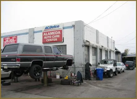  An SUV is lifted for maintenance outside Alameda Auto Care Center, with additional vehicles parked nearby and the building's signage visible.