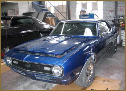  A blue classic car with its hood slightly open sits in a garage, surrounded by various tools and workshop equipment.