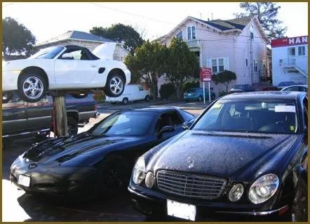  A white car on a lift, alongside a black car, at an auto care center parking area, with houses in the background.