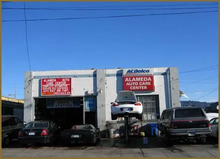  An auto repair shop with a car on a lift outside. Several other vehicles are parked in front of the building marked with "Alameda Auto Care Center" signs.