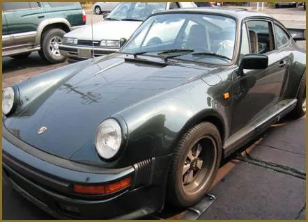  A dark-colored vintage sports car parked outside with visible reflections on its surface. Nearby are other parked vehicles.