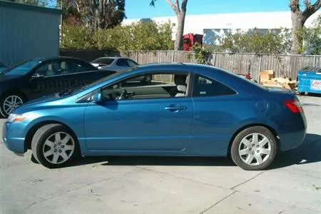 A blue two-door car parked outside on a concrete surface, surrounded by trees and other vehicles in the background.