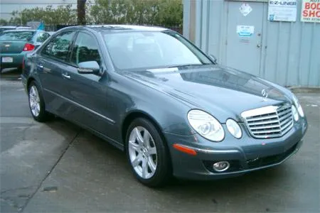  A shiny, restored gray sedan is parked on a wet concrete surface near an auto body shop's exterior wall.
