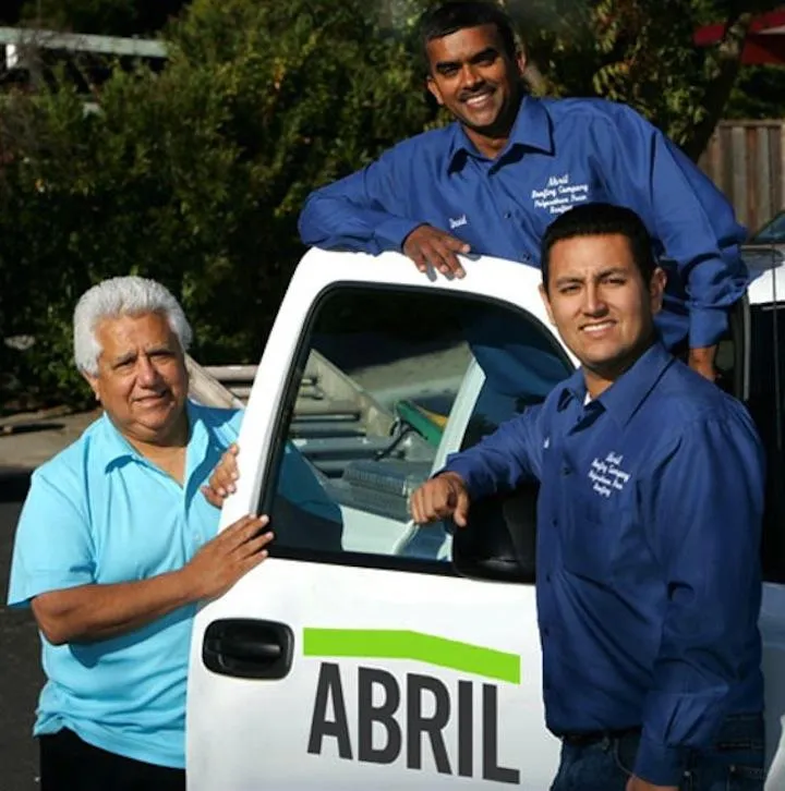 Three people wearing blue shirts stand by a vehicle with the Abril logo. Two are posing beside the door, while one leans on the roof. The background shows greenery.