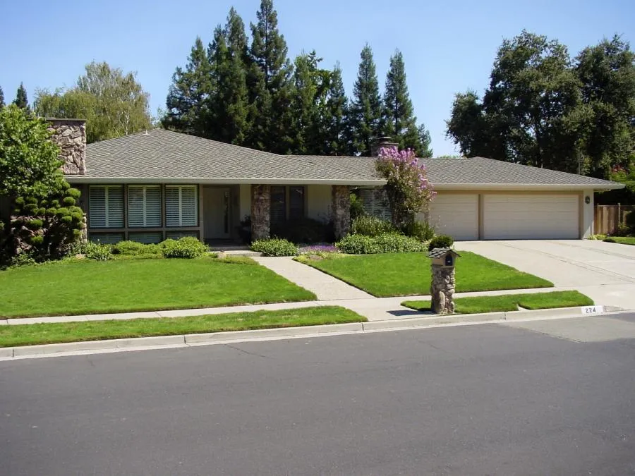  Single-story house with a clean roof, manicured lawn, and large trees in the background.