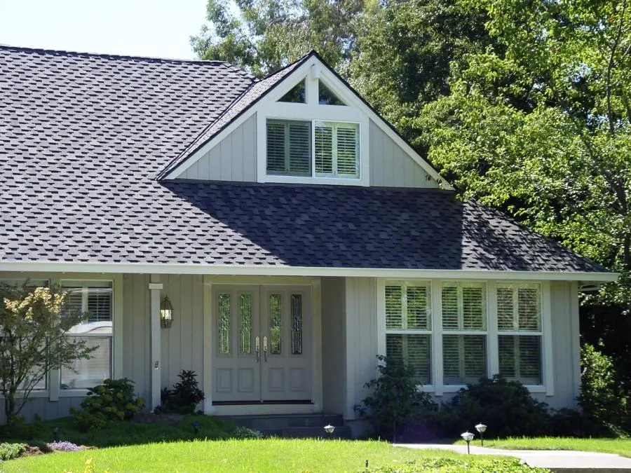  A house with a new shingle roof and dormer windows, surrounded by lush greenery, with sunlight highlighting the craftsmanship.