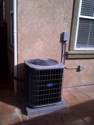 Outdoor air conditioning unit installed on concrete pad beside a residential home, near electrical panel and window.