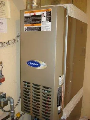  A Carrier heater installed in a room, showing various labels and warning stickers on its metallic surface. Visible pipes are connected to the unit.